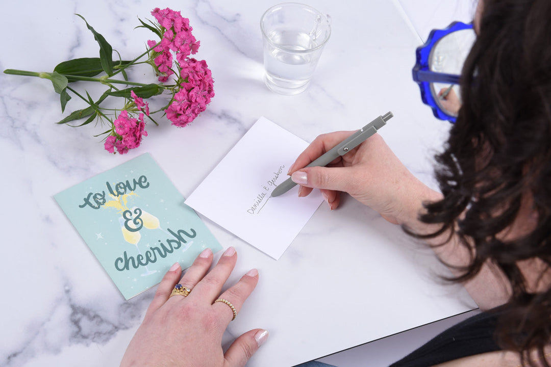 A woman addressing an envelope, and a greeting card that says "to love and cheerish" with an illustration of two champagne flutes clinking